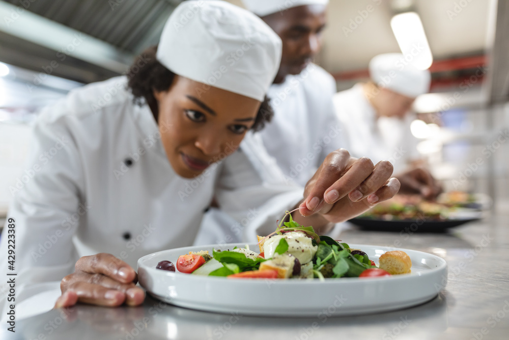 Mixed race professional chef finishing dish before serving with colleague in background Mixed race professional chef finishing dish before serving with colleague in background