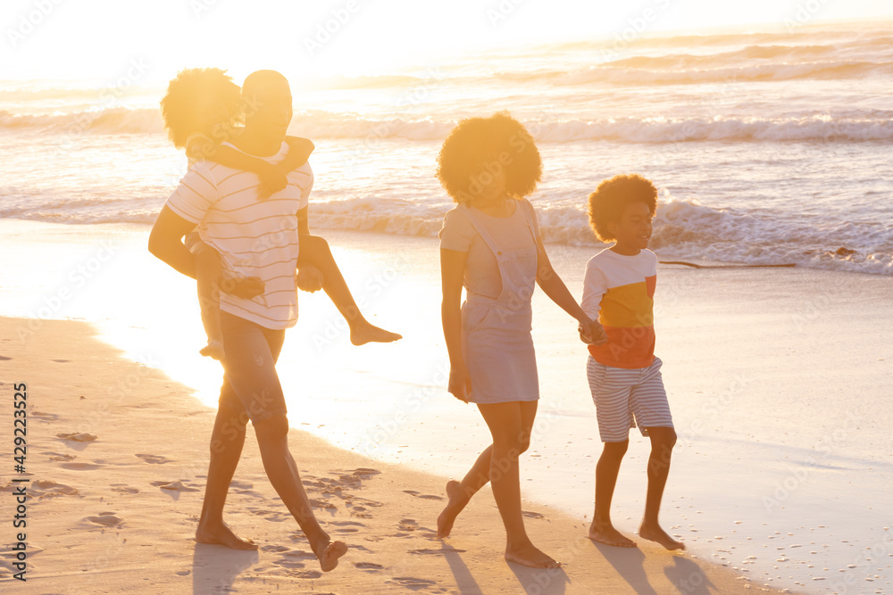 African american parents and two children walking holding hands and piggy backing at the beach