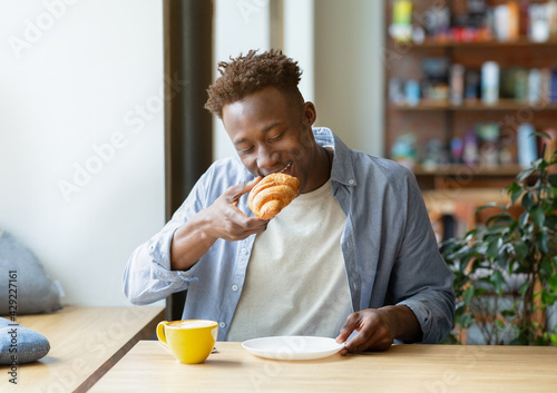 Cool Funky African American guy eating croissant with coffee, having breakfast at city cafe