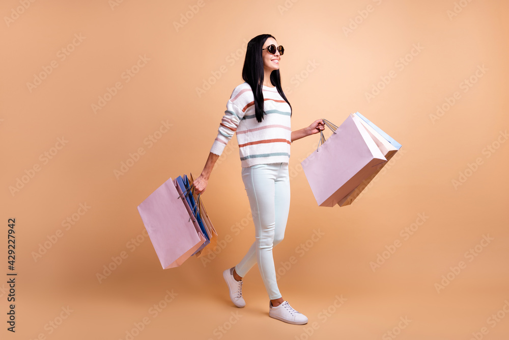 Full size profile side photo of young happy positive girl on shopping wear sunglasses isolated on beige color background Full size profile side photo of young happy positive girl on shopping wear sunglasses isolated on beige color background