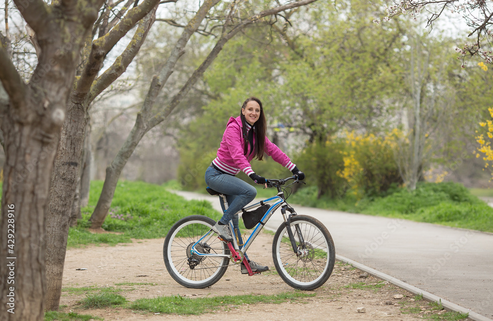 beautiful athletic girl riding a bike, wearing jeans and a pink jacket, spring landscape