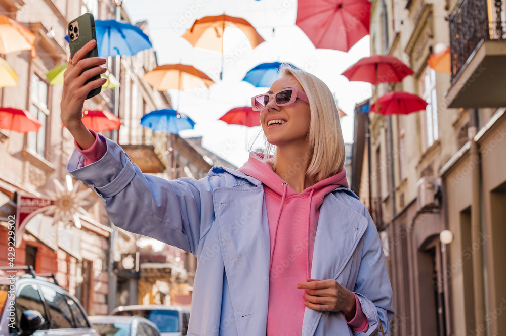Obraz premium Happy smiling fashionable woman makes selfie with her smart phone in street of European city