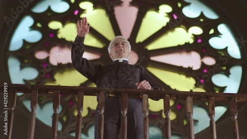 Low angle view of aged Caucasian clergyman wearing black clothes and white collar standing in Christian church against stained-glass window and preaching Sunday sermon