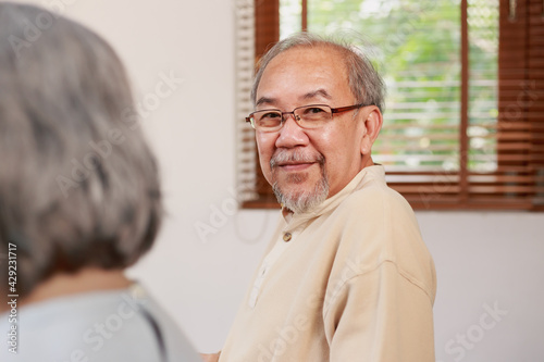 Happy Asian senior man sitting by white hair elderly woman looking at camera with smiley face by the window at home. Friendly Asian retired people with beard wearing glasses with happy smile indoor