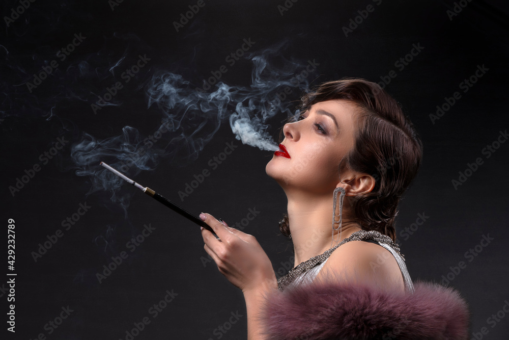 Obraz premium Retro woman portrait. A beautiful woman in the style of the 20s and 30s with a cigarette holder smokes.