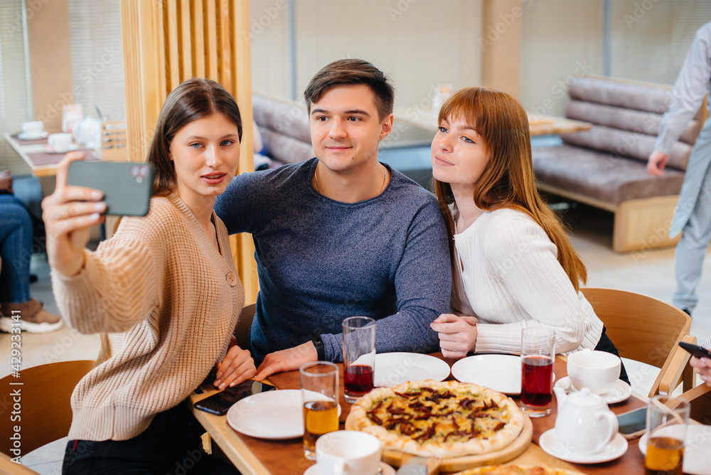 A group of young cheerful friends is sitting in a cafe talking and taking selfies on the phone. Lunch at the pizzeria.