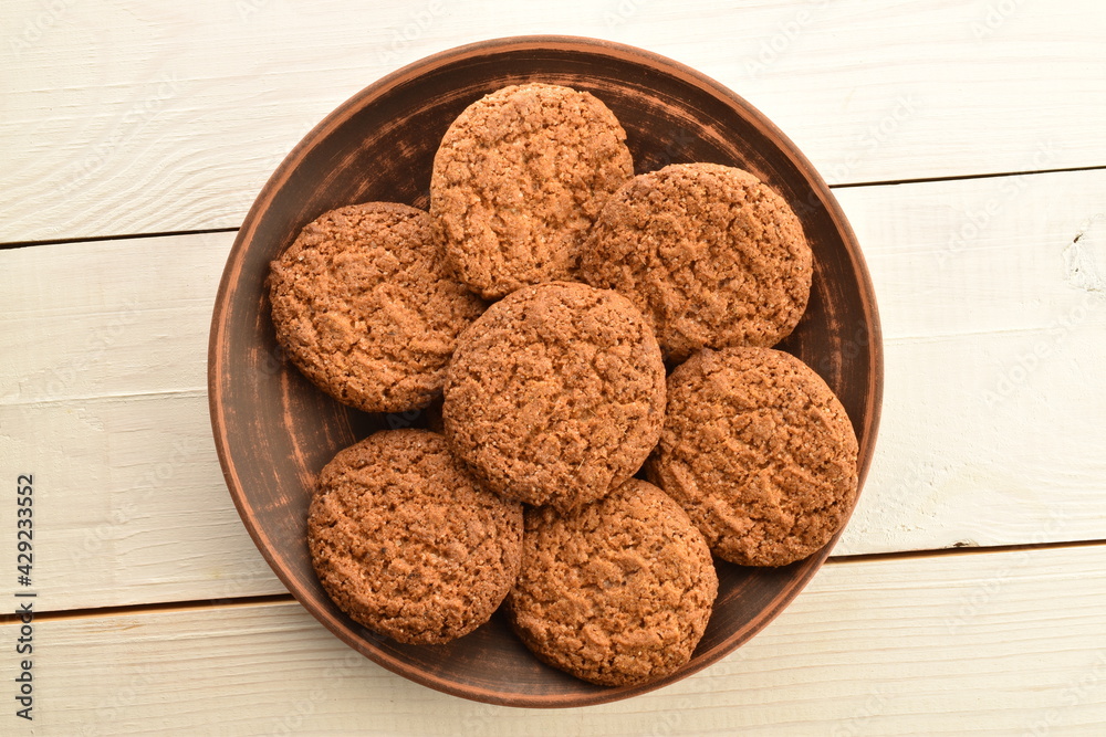 Several dark brown oatmeal cookies in an earthenware dish on a wooden table, close-up, top view. Several dark brown oatmeal cookies in an earthenware dish on a wooden table, close-up, top view.