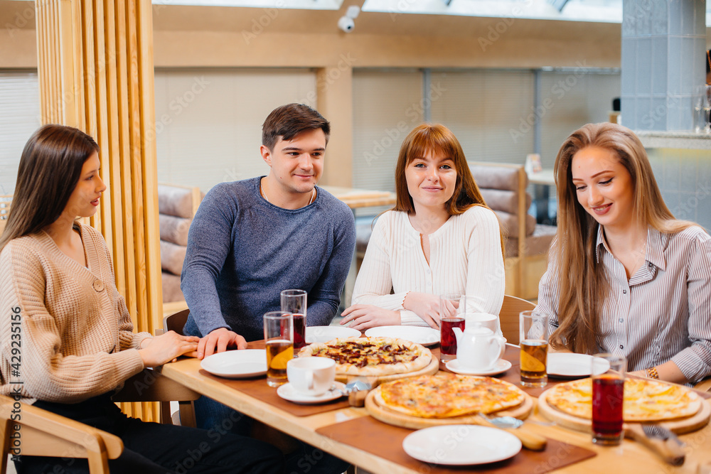 A group of young cheerful friends is sitting in a cafe talking and eating pizza. Lunch at the pizzeria. A group of young cheerful friends is sitting in a cafe talking and eating pizza. Lunch at the pizzeria.