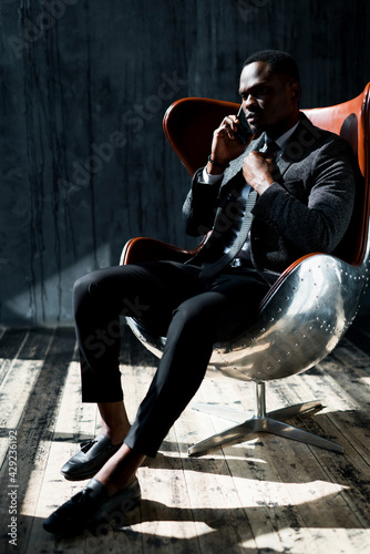 African American director of the company sits in a chair in a suit and talks on the phone © DmitryStock