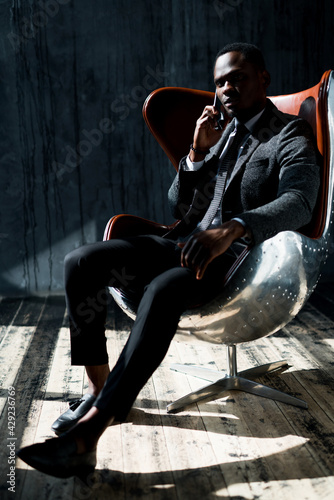 African American director of the company sits in a chair in a suit and talks on the phone © DmitryStock