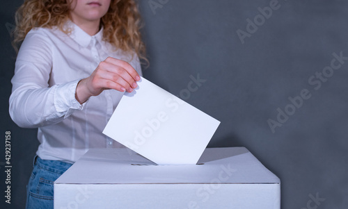 Junge Frau mit lockigem Haar  im weissen Hemd steht hinter einer Wahlbox und wirft ihren Stimmzettel hinein.  photo