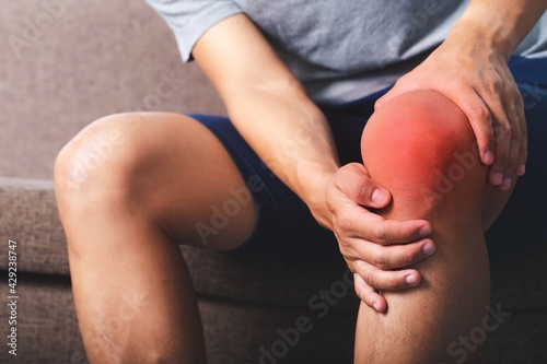 Image of a young man sitting on sofa holding his knees in pain.,Patellar tendon Inflammation.Healthcare and medical concept.