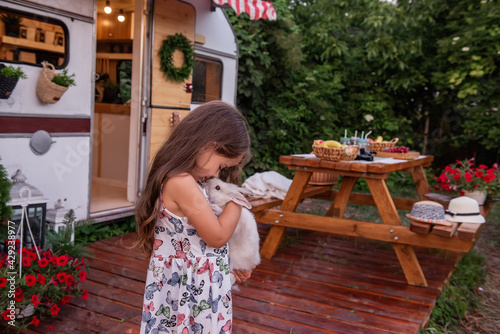 Canvas Print Portrait of a little girl in a straw hat sits near a trailer truck at home on a wooden floor, holding a fluffy rabbit in her hands