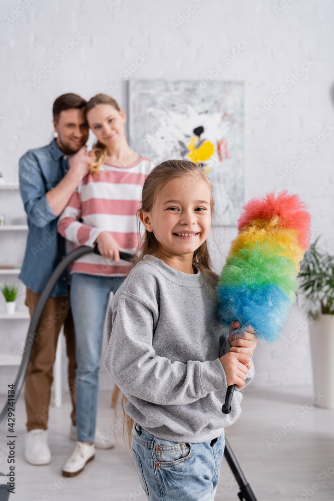 Positive kid holding dust brush near parents with vacuum cleaner on blurred background.