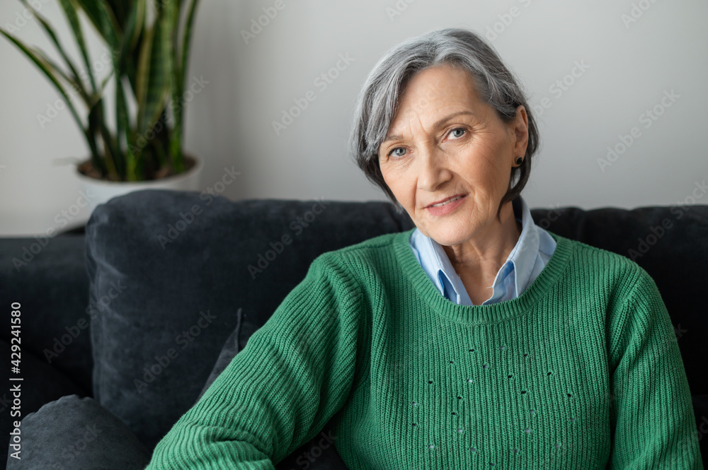 A portrait of a charming beaming old lady posing and looking at the camera,  gray-haired senior mature woman sitting in the living room, wearing a  casual green jumper, resting and smiling foto