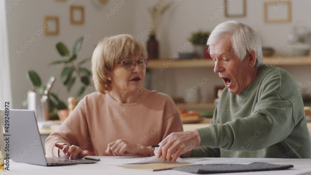 Senior couple sitting at table, using laptop, going through financial papers and discussing family budget at home Senior couple sitting at table, using laptop, going through financial papers and discussing family budget at home