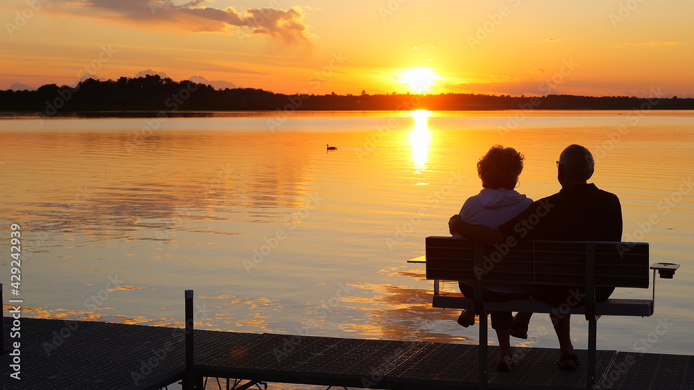 Silhouette of two people on a bench on a dock as they enjoy a sunset over a beautiful lake in Minnesota on a serene and relaxing evening, while a Canada goose swims by on the water. Silhouette of two people on a bench on a dock as they enjoy a sunset over a beautiful lake in Minnesota on a serene and relaxing evening, while a Canada goose swims by on the water.