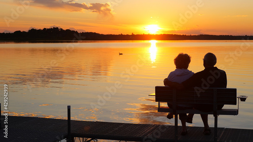 Silhouette of two people on a bench on a dock as they enjoy a sunset over a beautiful lake in Minnesota on a serene and relaxing evening, while a Canada goose swims by on the water. © scandamerican