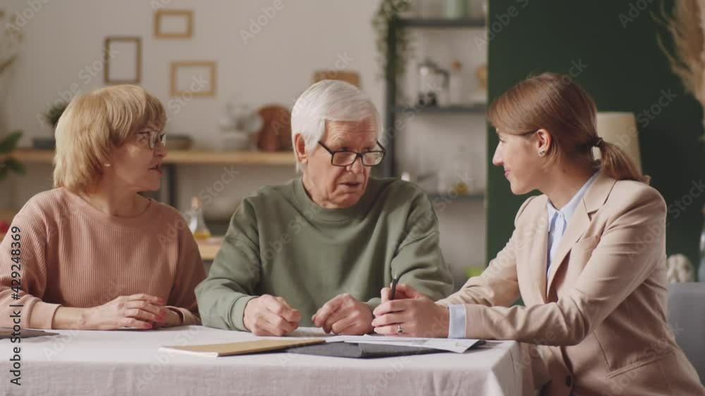 Elderly couple and female financial advisor having discussion, then looking together at camera and smiling while sitting at table at home