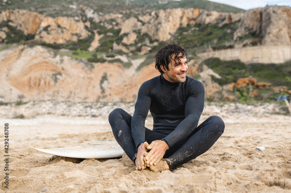 Young surfer man wearing diving suit sitting on the sand next to his surfboard and resting after surfing. Young surfer man wearing diving suit sitting on the sand next to his surfboard and resting after surfing.
