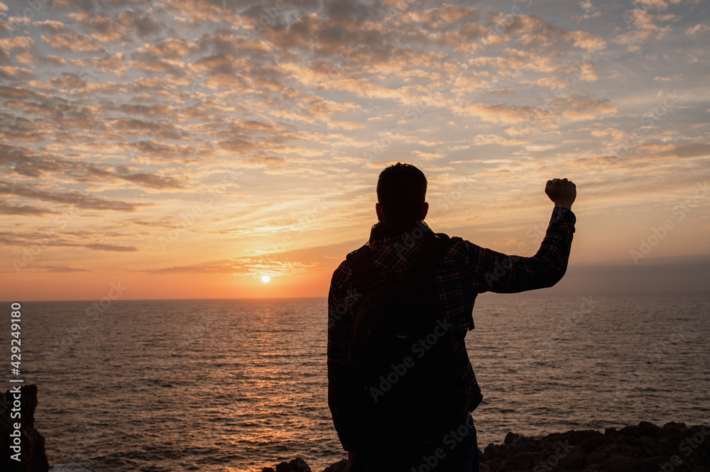 Back view silhouette of unrecognizable man standing against the sea looking sunset raising arms and showing victory fist.