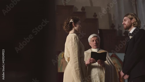 Tracking right of young Caucasian man and his charming bride standing at altar in Lutheran church and pronouncing wedding vows while senior priest reading Bible photo