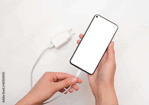 Connecting charging a smartphone via a USB cable and a power supply unit on a white background, hand connects. Smartphone with a white screen. Concept of rechargeable smartphone via USB cable.top view