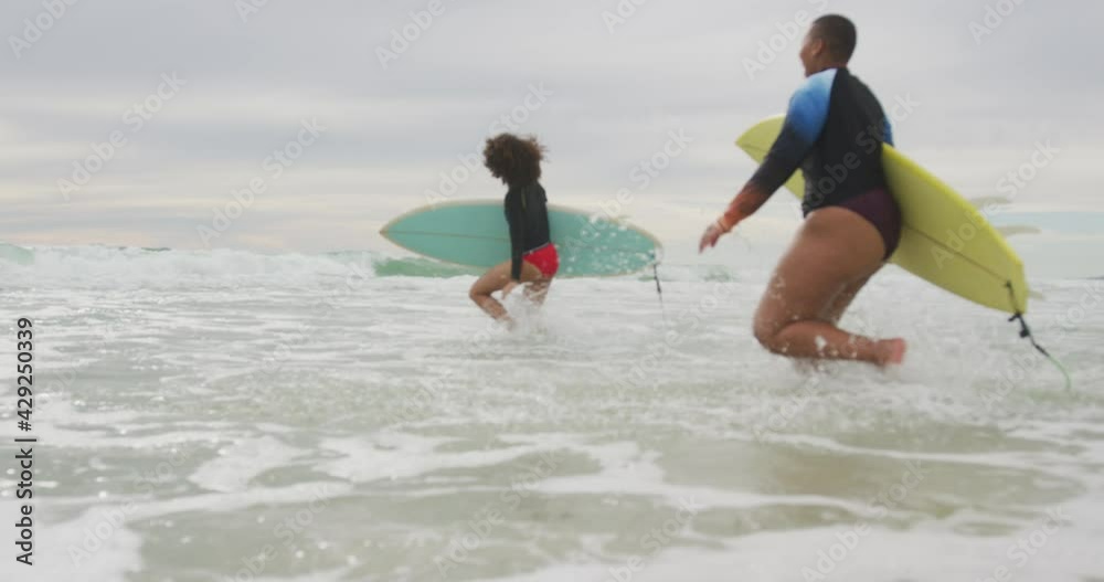 Happy african american female friends running into the sea holding surfboards Happy african american female friends running into the sea holding surfboards