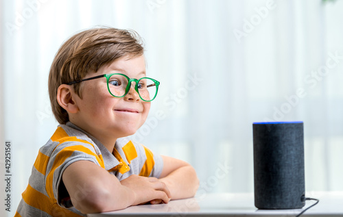 Children, education and technology concept. Funny pupil boy using smart speaker at home while doing homework. Home robot. Education programme for child.