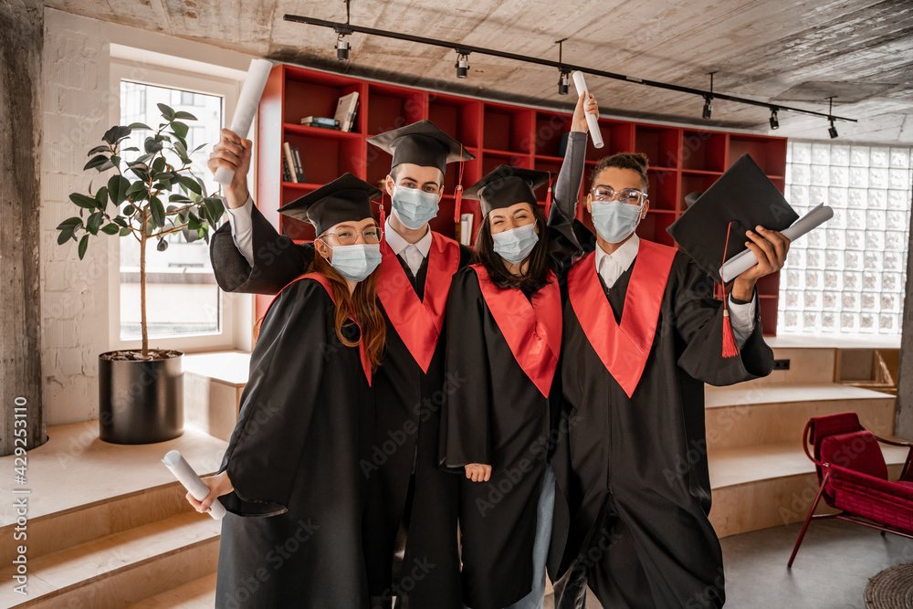 multiethnic students in medical masks, gowns and caps holding diploma, graduation class 2021