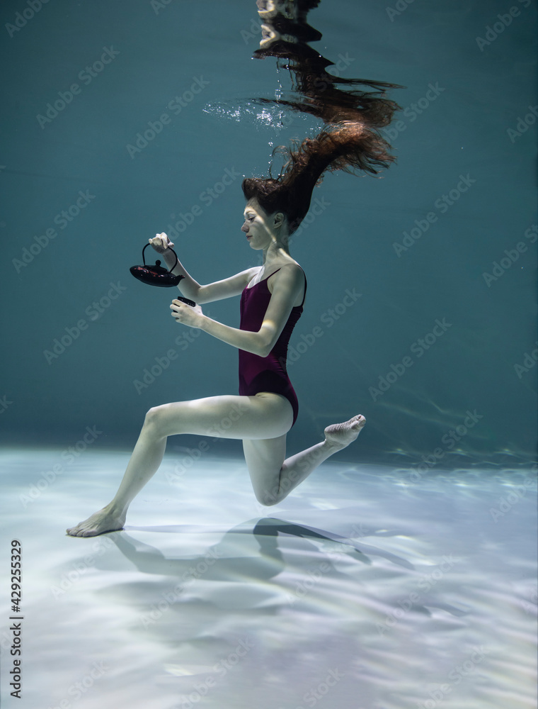 beautiful girl in a red swimsuit with a colored teapot and a cup is having a tea party under water on a blue background