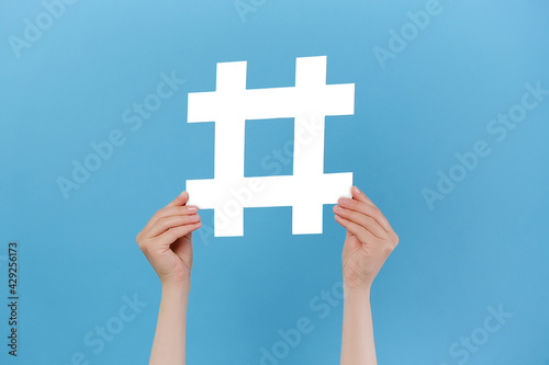 Horizontal images of female hands holding large big white hashtag sign, viral web content, internet promotion, isolated on blue studio background. Concept of trendy social media posts and blogging