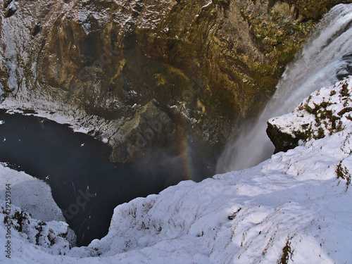 High angle view of famous cascade Skógafoss (height 60m) falling down a steep rocky slope near the southern coast of Iceland with colorful rainbow on sunny winter day with snow.