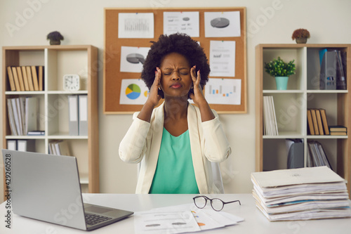 Young badly unwell stressed overwhelmed african american businesswoman rubbing temples feeling exhausted suffering from headache sitting at office desk portrait. Stress at work or migraine concept photo