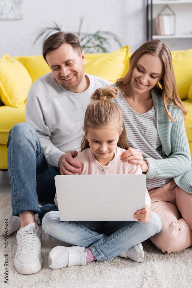 Child using laptop near smiling parents on blurred background at home. Child using laptop near smiling parents on blurred background at home.