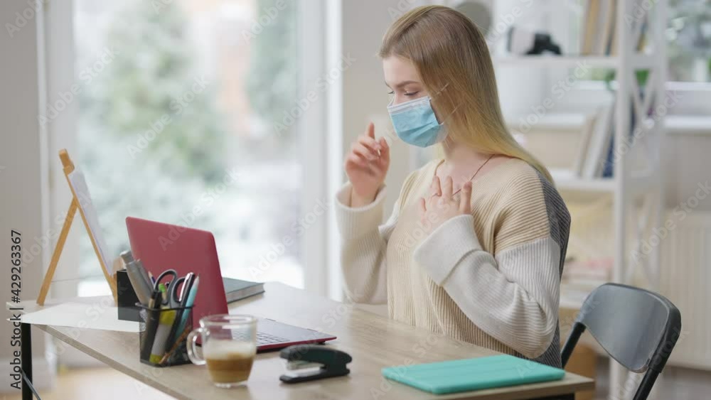 Focused slim young woman in Covid-19 face mask using sign language talking in video chat at home. Portrait of confident Caucasian lady chatting online with deaf person on coronavirus pandemic