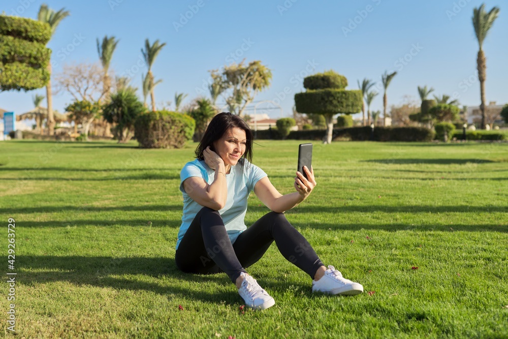 Mature woman doing morning exercises on green grass in park, with bottle of water