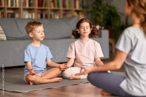 Children brother and sister sitting in lotus pose on floor and meditating with closed eyes with mom