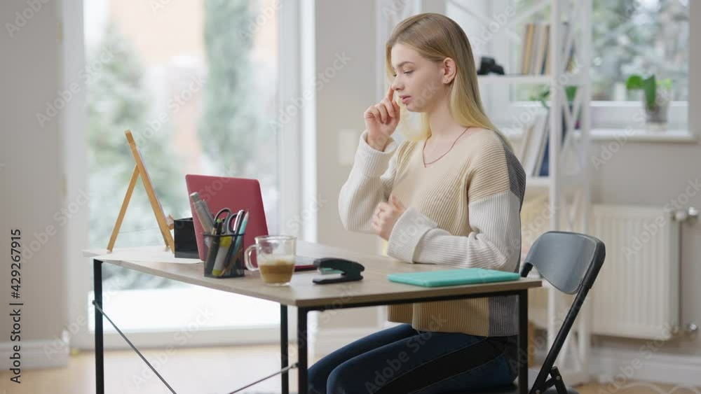 Positive smiling young woman waving in video chat and gesturing using sign language. Side view portrait of cheerful charming Caucasian lady communicating with deaf person online at home