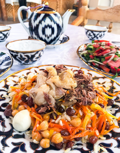 A set table in an Uzbek restaurant: wedding pilaf made of rice with carrots. With lamb and tail fat. As well raisins, chickpeas and eggs. On blurry background, a tomato salad and a teapot with bowls