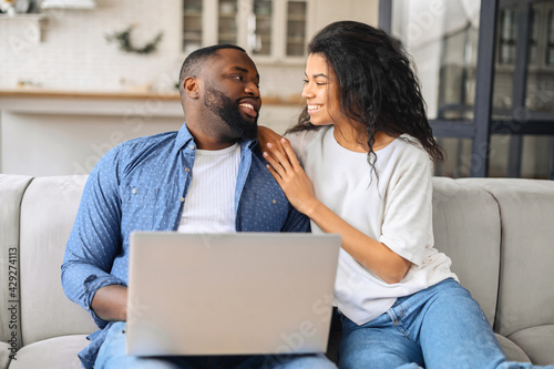 Cute African-American couple in love spends leisure together with the laptop at home, boyfriend and girlfriend sitting on the couch in embraces and looks at each other tenderly