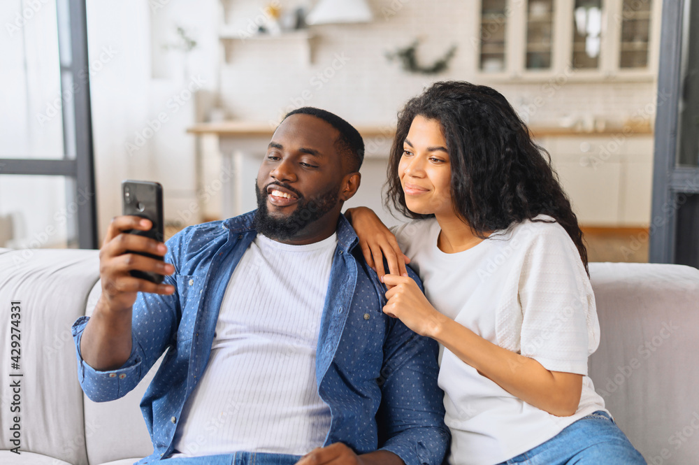 Obraz premium African-American couple with smartphone on the couch spends leisure time in social networks, scrolling feed, watching videos, a black guy is showing smth to his girlfriend