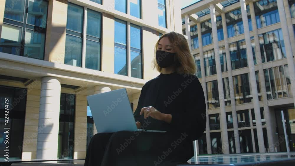 Asian girl works on laptop outdoors during covid-19 coronavirus pandemic. Female manager or designer wearing black clothing and protective mask sits outside and uses computer Asian girl works on laptop outdoors during covid-19 coronavirus pandemic. Female manager or designer wearing black clothing and protective mask sits outside and uses computer