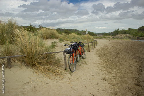 Fototapeta Naklejka Na Ścianę i Meble -  Two orange bicycles laden with backpacks leaning against a beach resort with the dune background. Bicycle touring concept. Castiglione della Pescaia, Tuscany