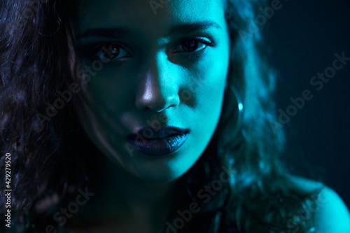 Close up portrait of attractive woman with trendy accessories and with curly hair looking at camera on dark blue light. Concept of professional shooting for glamour magazine. 