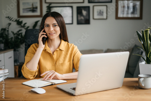 Young woman talking on mobile phone while working with laptop at desk. Home office