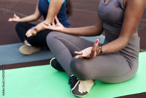 Close frame of hands in mudra gesture of women doing yoga outdoors. Caucasian and black woman sitting in lotus position in the stadium