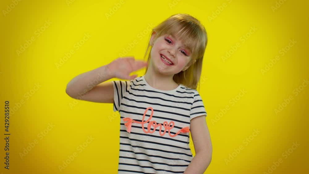 Little blonde child kid girl waves hand palm in hello gesture welcomes someone with hospitable expression expresses positive emotions. Yellow studio background. Childhood lifestyle. Young children Little blonde child kid girl waves hand palm in hello gesture welcomes someone with hospitable expression expresses positive emotions. Yellow studio background. Childhood lifestyle. Young children
