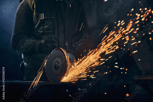 Close up of man in special dark suit and black protective gloves working angle grinder for metal with large flash sparks. Concept of process working electric steel cutter machine. photo