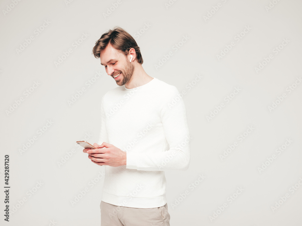 Handsome smiling hipster lumbersexual businessman model wearing white casual sweater and trousers.Fashion stylish man posing against gray background in studio. Holding smartphone in hands.In earphones Handsome smiling hipster lumbersexual businessman model wearing white casual sweater and trousers.Fashion stylish man posing against gray background in studio. Holding smartphone in hands.In earphones